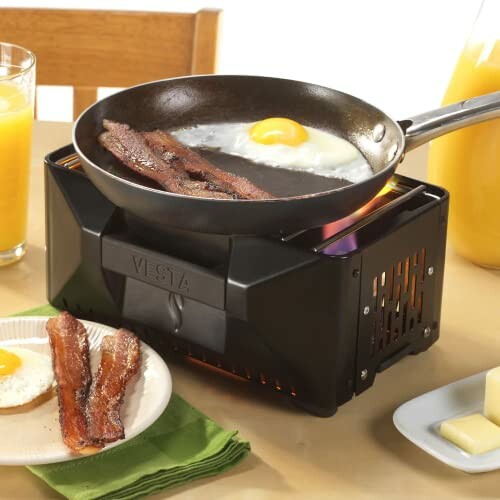 Portable stove cooking bacon and eggs with juice and toast.